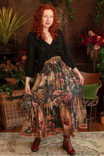 Load image into Gallery viewer, Woman wearing a black top and floral skirt in a decorative indoor setting with plants and a wooden wall.