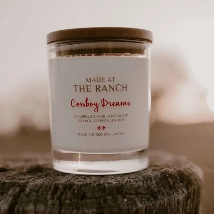 Candle labeled 'Made at the Ranch Cowboy Dreams' on a wooden surface with a blurred background