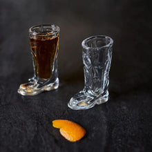 Load image into Gallery viewer, Two glass boot-shaped shot glasses with a dark liquid and an orange peel on a black background