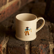 Load image into Gallery viewer, A cream-colored ceramic mug with a small hand-painted cowboy figure, resting on a rustic brick surface.