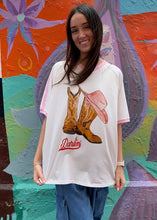 Load image into Gallery viewer, Woman wearing a white t-shirt with cowboy boots and a pink cowgirl hat design, standing in front of a colourful mural.
