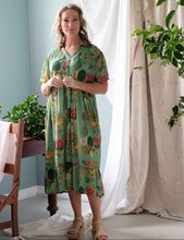 Load image into Gallery viewer, Woman wearing a green floral dress standing in a room with light blue walls and white curtains.
