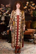 Load image into Gallery viewer, Woman in a patterned floral kimono standing in a decorated room with flowers and furniture.