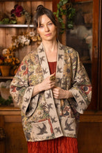 Load image into Gallery viewer, A woman stands in a rustic room with wooden cabinets and plants, wearing a beige kimono-style jacket with a bird pattern over a red top and a long red skirt.