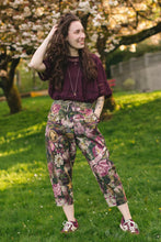 Load image into Gallery viewer, Woman in a purple top and floral pants standing in a park with cherry blossom trees.