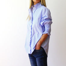 Load image into Gallery viewer, A woman wearing a light blue and white vertically striped button-up shirt with ruffled trim on the collar and cuffs, paired with dark blue jeans, shown from the side against a plain white background.